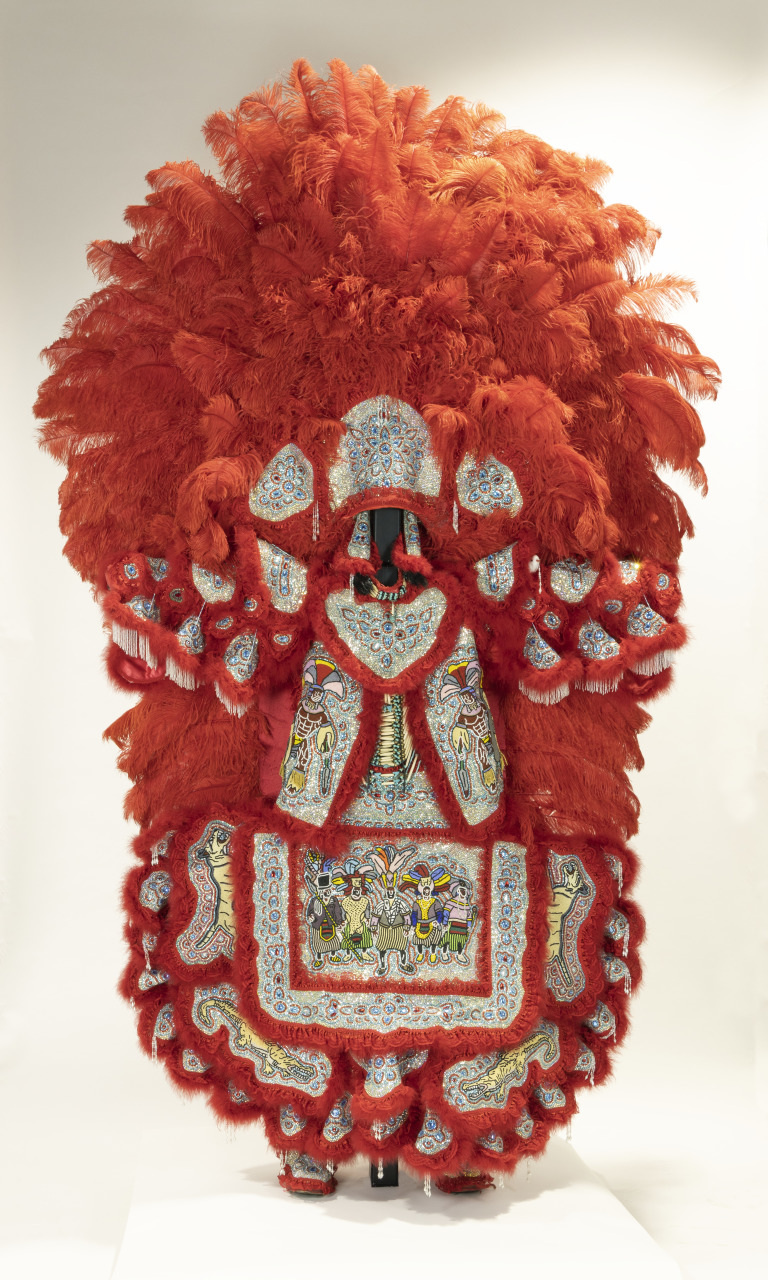 A person is wearing an elaborate costume adorned with large red feathers and intricate beaded embroidery depicting scenes and patterns. The costume covers the entire body and includes a headdress and multiple layered elements, creating a dramatic appearance.