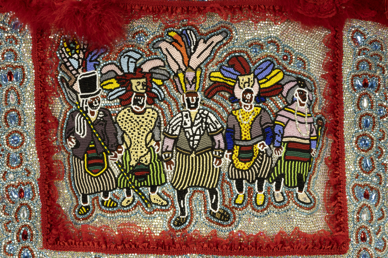 Beadwork depicting four stylized figures in traditional attire with vibrant colors and intricate designs. They are adorned with headdresses and surrounded by a red feathered border.