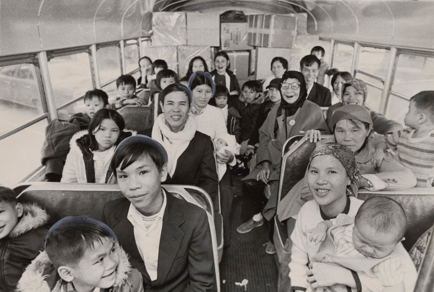 A black and white photo shows a crowded bus interior with smiling adults and children. Some wear scarves and hats. The bus is filled with passengers sitting close together, many looking towards the camera.