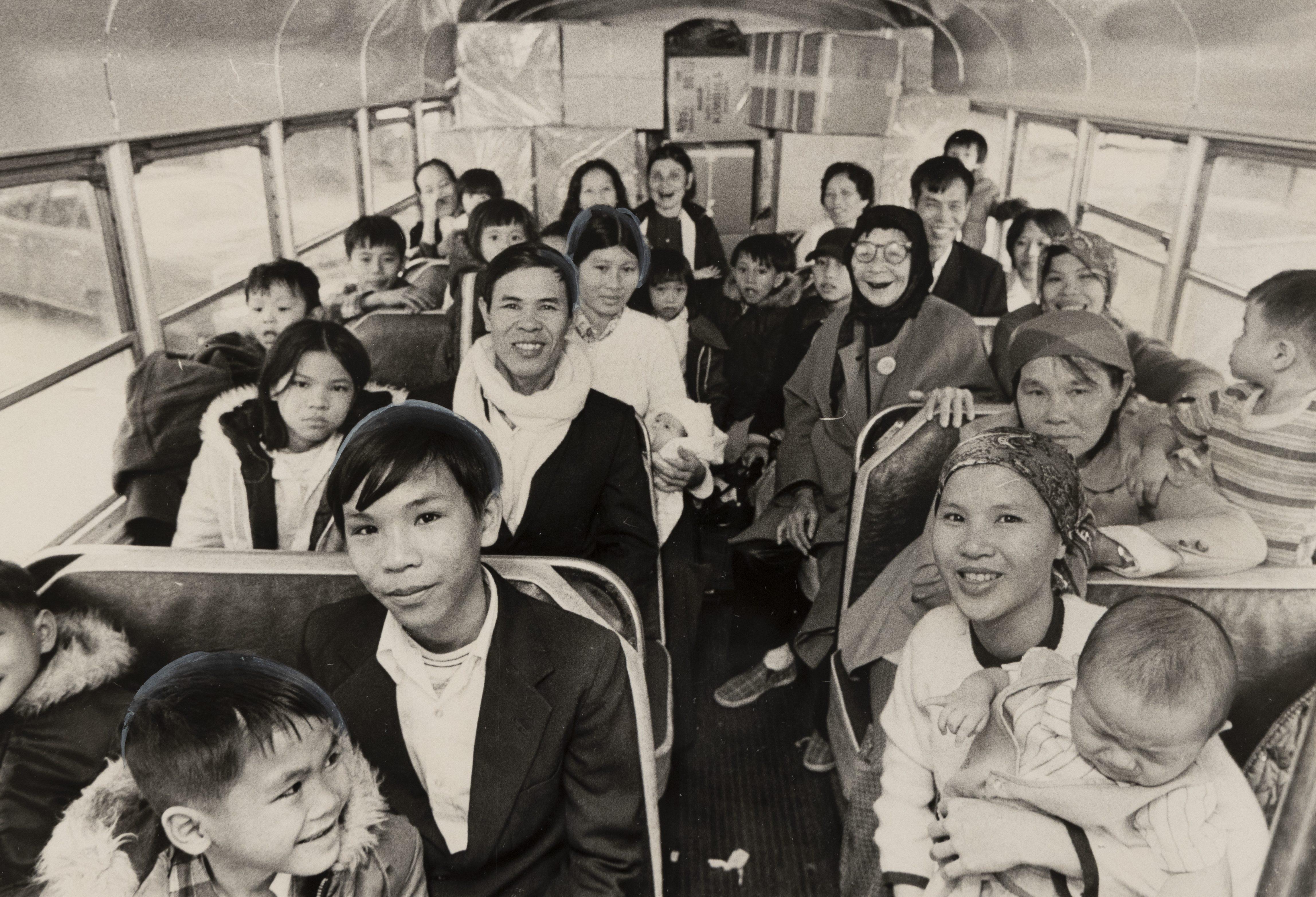A black-and-white photo of a diverse group of people, including men, women, and children, sitting inside a bus. They appear cheerful and relaxed, with some smiling at the camera. The bus is filled with people seated closely together.