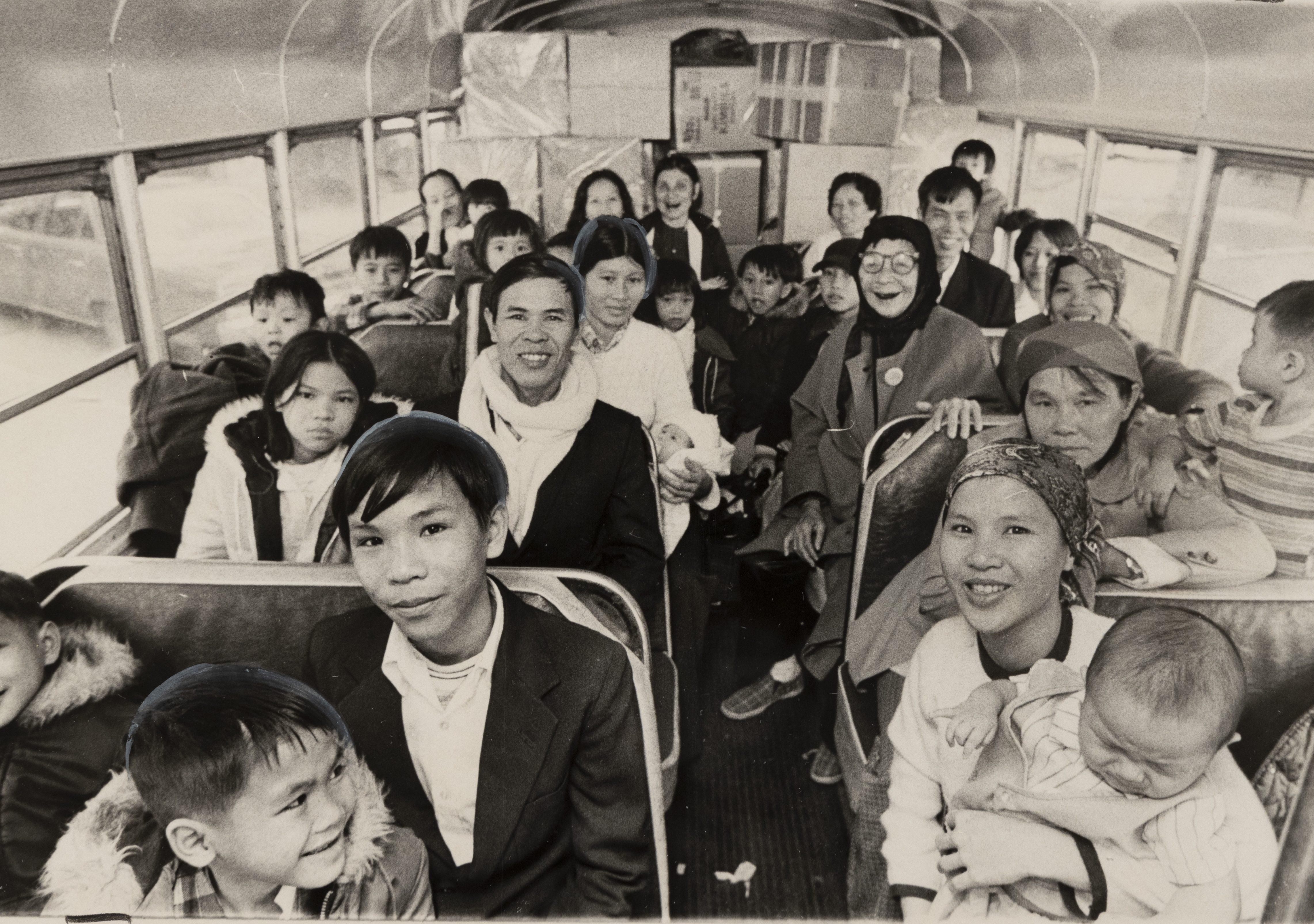 A black and white photo of a group of people, including adults and children, sitting closely together on a bus. Most are smiling and looking toward the camera. The interior of the bus is visible, with seats and windows on either side.