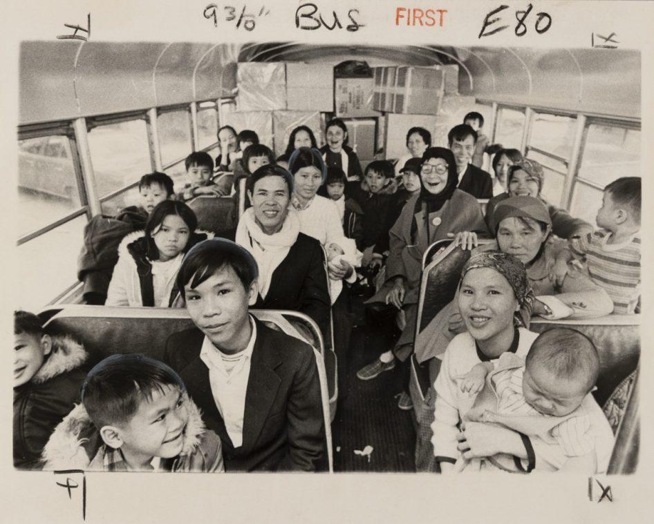 A vintage photo of a crowded bus interior with men, women, and children seated. The people are smiling and dressed in casual clothing. Handwritten notes are visible at the top of the image.