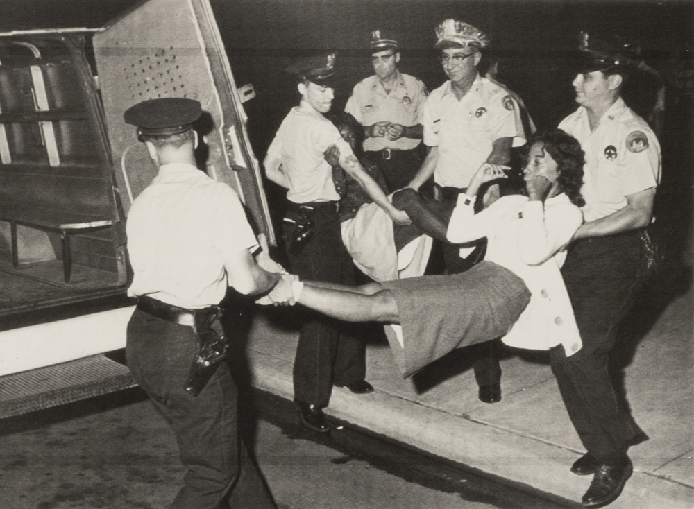 Black and white photo of a woman being carried by multiple police officers toward a police van. She is wearing a skirt and jacket, while the officers are in uniform. The scene is at night on a sidewalk.