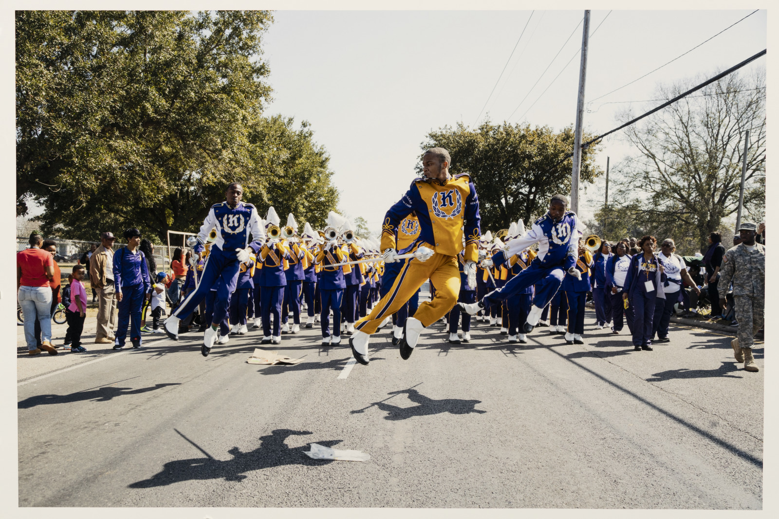 A marching band in vibrant blue and yellow uniforms performs energetically on a street. Band members play instruments while two dancers leap in synchronization. Spectators line the street, enjoying the sunny day and lively performance.