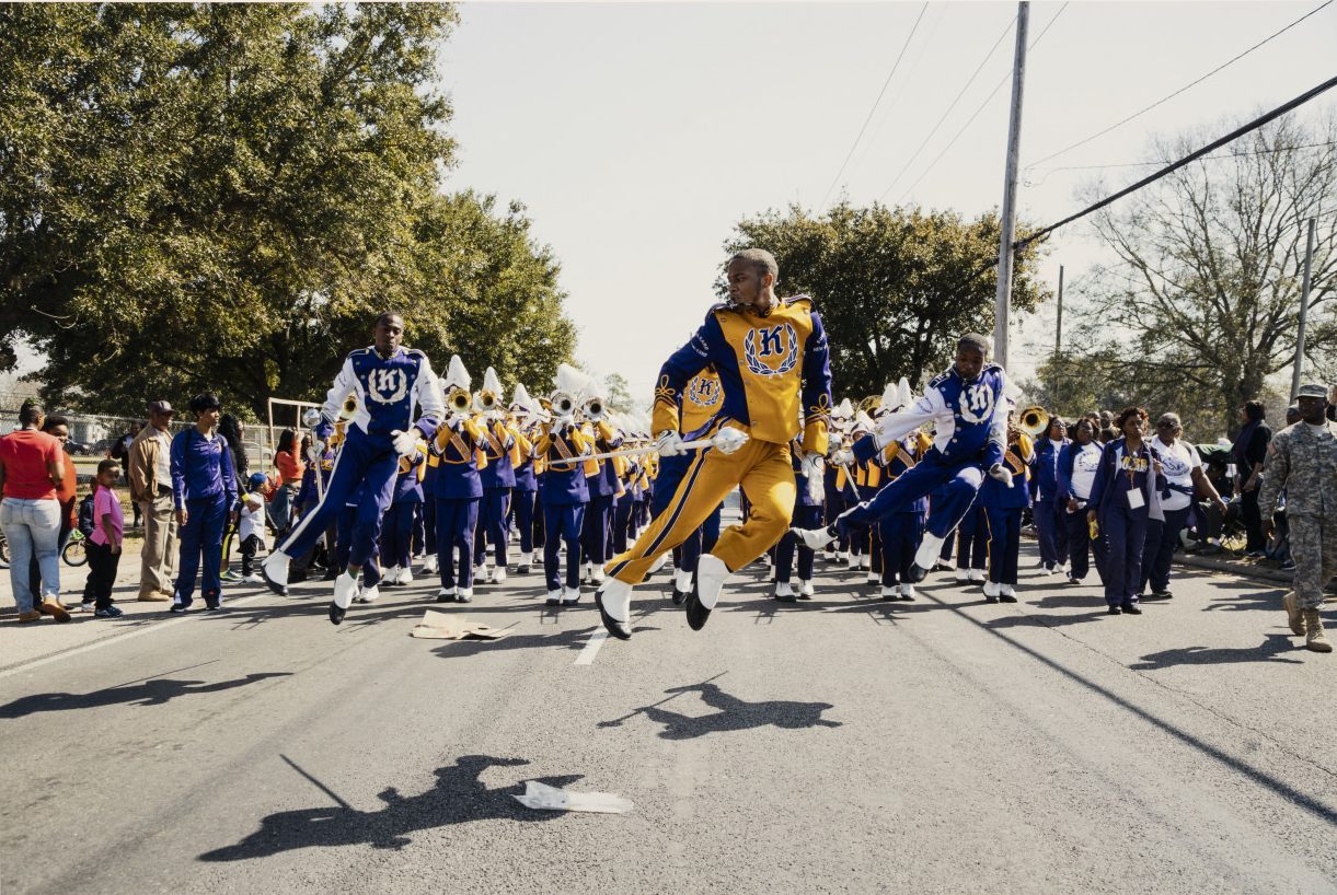 Marching band members in blue and yellow uniforms perform energetically down a street. Some are jumping mid-air, with instruments raised, while spectators watch from the sidelines. Trees and a clear sky form the backdrop.