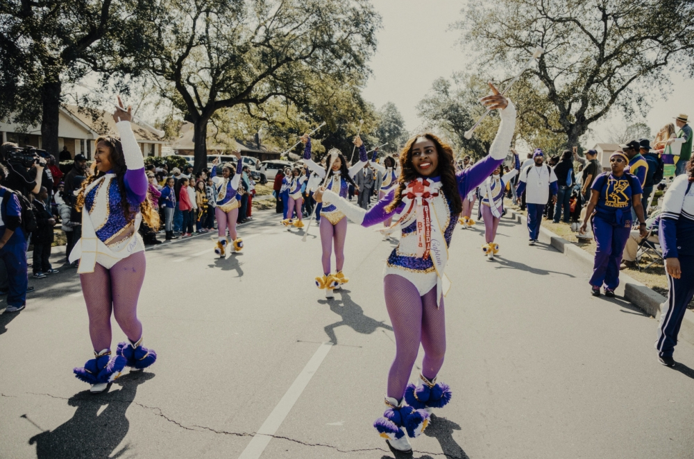 A group of dancers in purple and white costumes performs energetically in a parade. They are surrounded by onlookers on a tree-lined street, with some people playing instruments in the background.