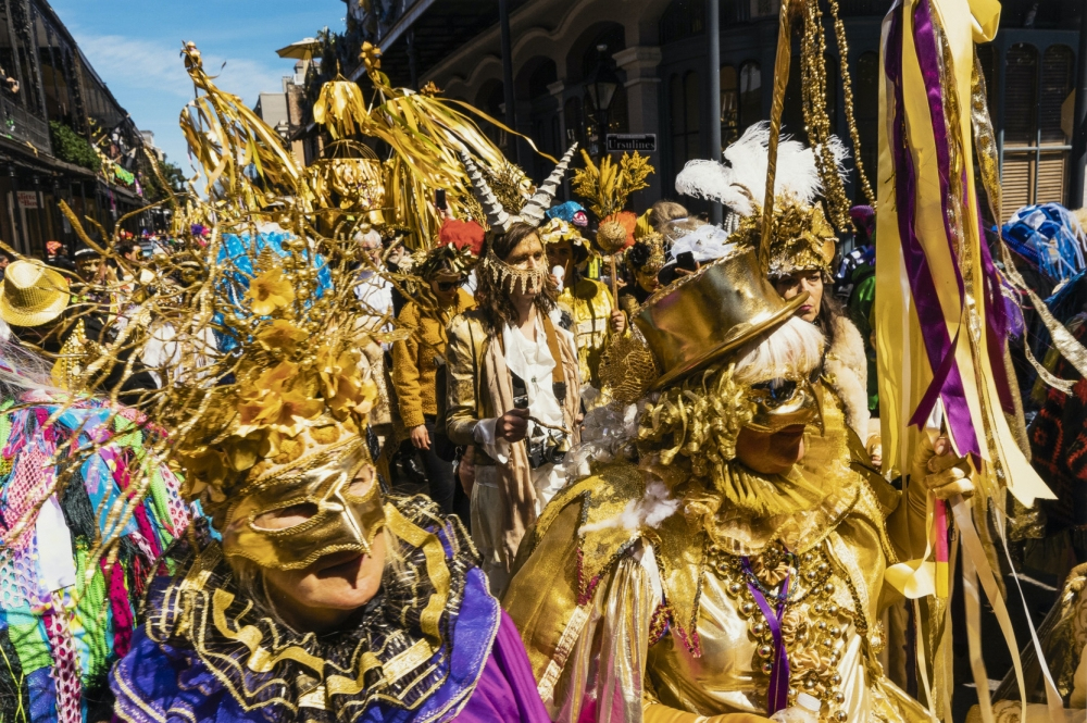 A vibrant parade features individuals dressed in elaborate golden costumes and masks, adorned with feathers and ribbons. The lively scene captures the festive atmosphere of a cultural celebration in a bustling street setting.