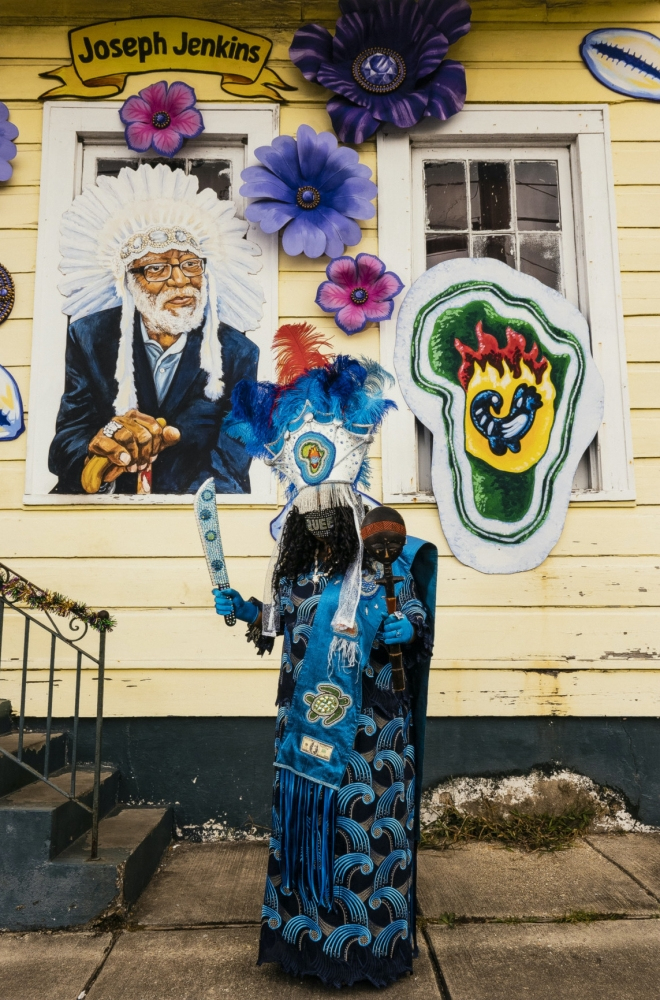 A person dressed in vibrant blue and white attire with feathers stands in front of a yellow building. The building features murals, including a portrait of an older man in a headdress and colorful designs. A sign above reads Joseph Jenkins.