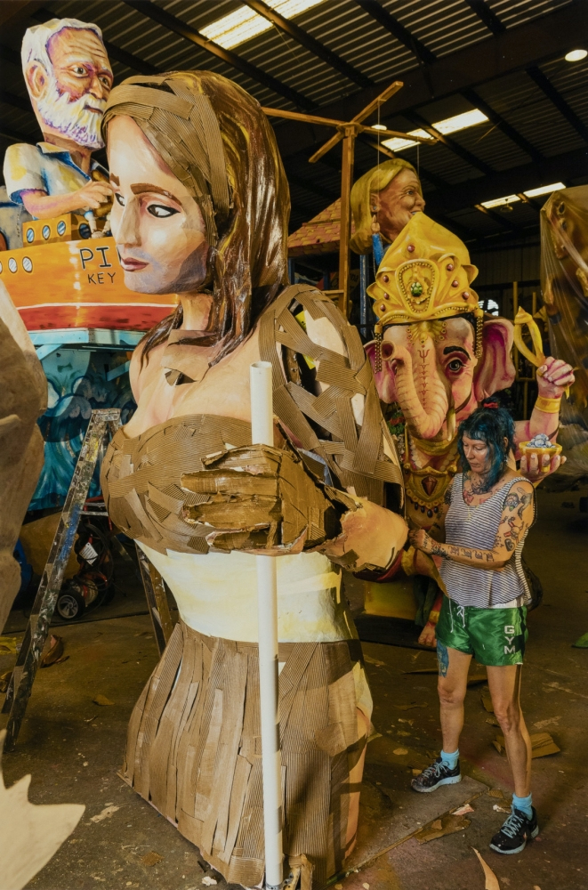 A person works on a large cardboard sculpture of a woman, surrounded by other colorful sculptures, including a figure with an elephant head wearing a crown, inside a warehouse with a high ceiling.