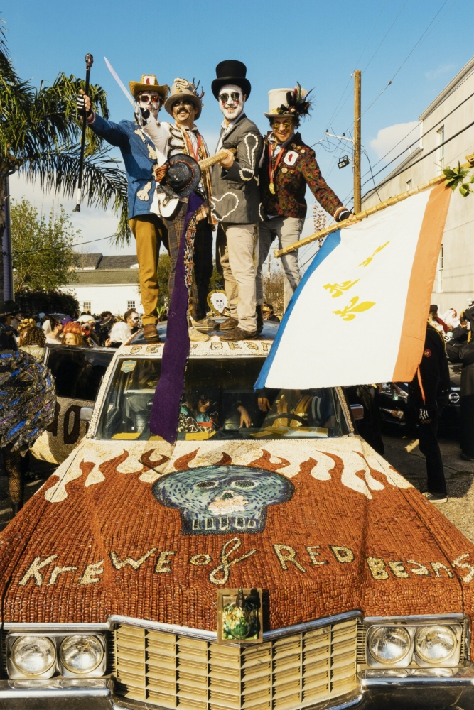 A group of people in colorful costumes and face paint stand on an elaborately decorated car. The car features a skull motif and the words Krewe of Red Beans. They appear to be part of a festive parade.