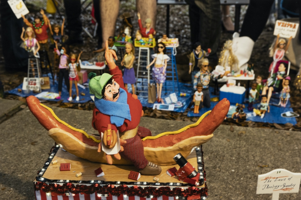 A whimsical sculpture of a joyful figure riding a hot dog bun like a rodeo, surrounded by miniature figures and scenes on a table. The background shows more small figures and tiny settings, adding a playful and creative atmosphere.