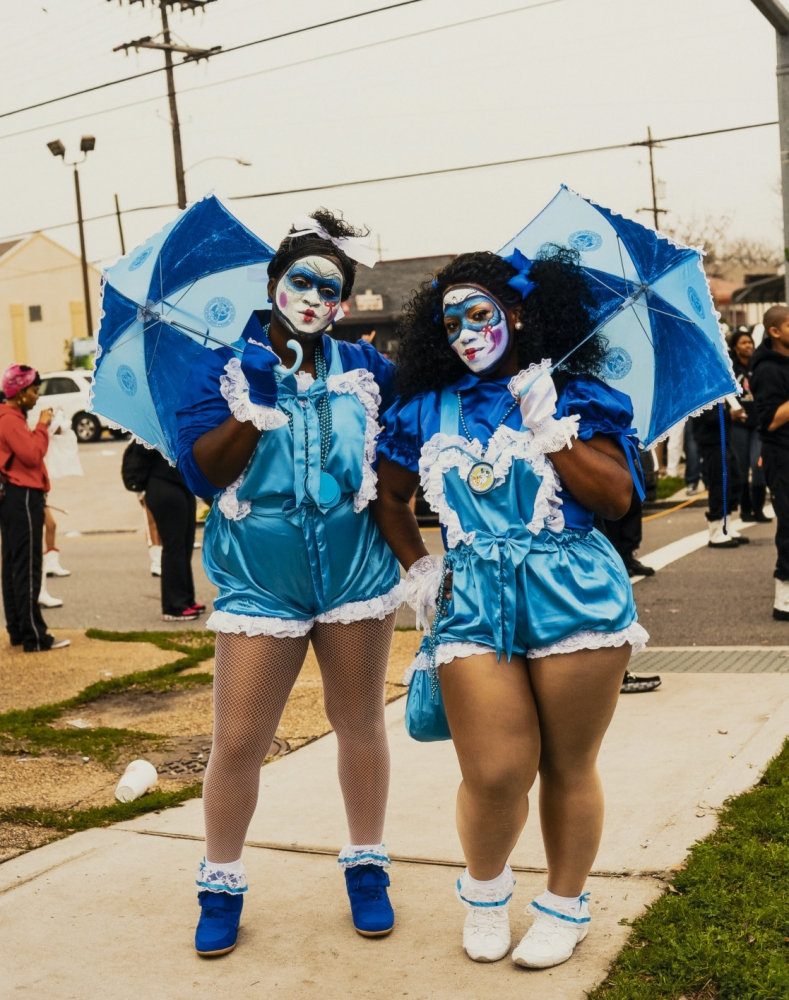 Two people dressed in vibrant blue costumes with white lace and clown-like face paint stand holding matching blue umbrellas. They appear to be part of a festive parade or event, with others visible in the background.