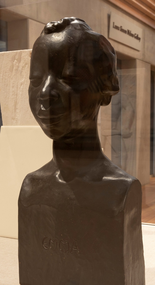A bronze sculpture of a persons upper body and head on display in a museum. The artwork features detailed facial features and the name Emma inscribed on the front. The background shows a gallery setting with minimal decor.