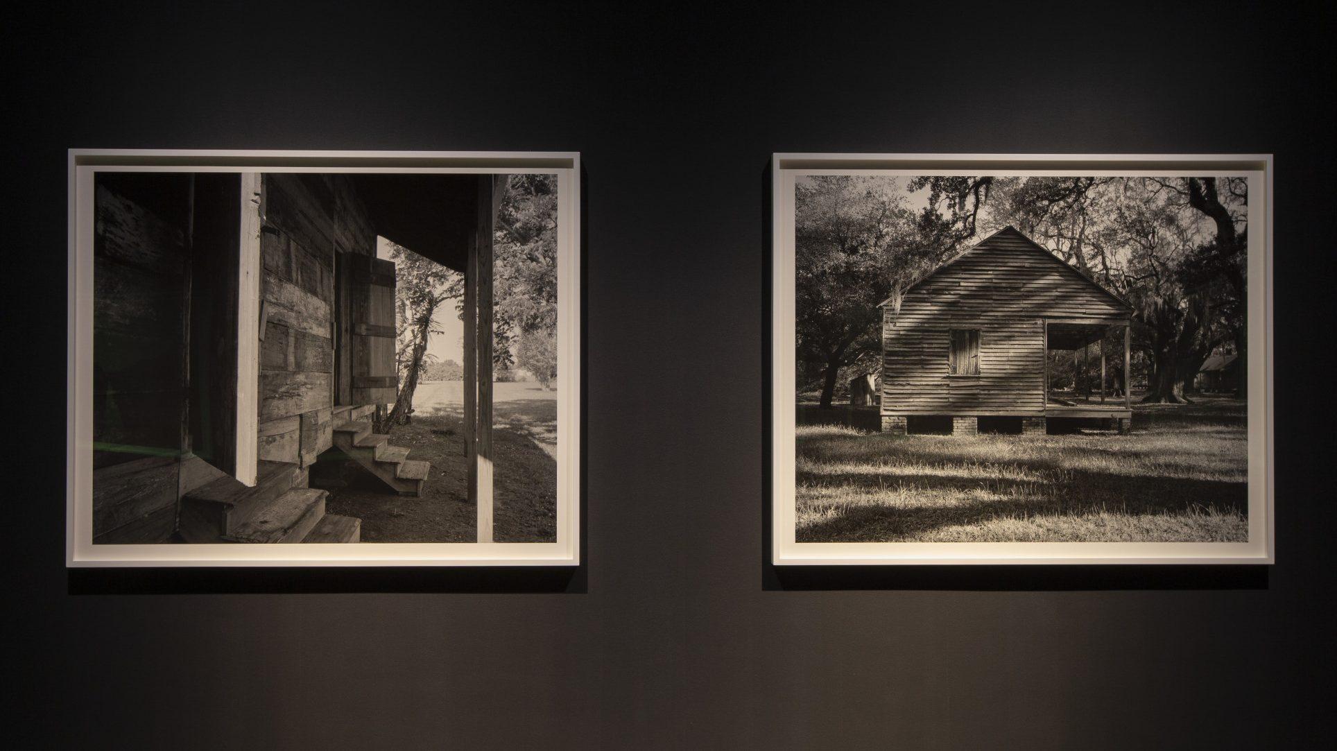 Two black-and-white framed photographs displayed on a dark wall. The left image shows a wooden porch with steps, while the right image features a small wooden house with trees in the background. Both evoke a rustic, serene atmosphere.