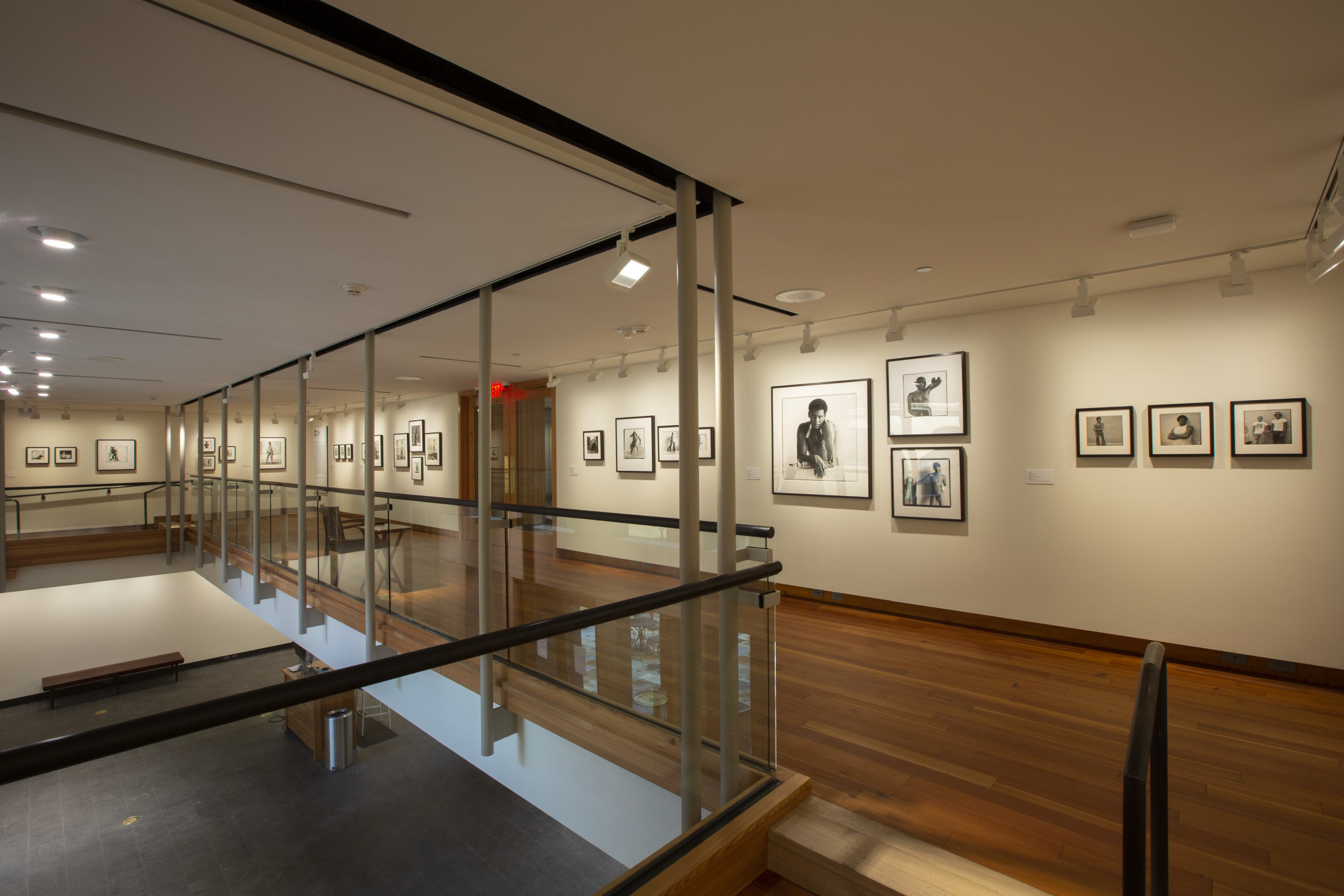 A modern art gallery with wooden floors and beige walls displays framed black-and-white photographs. The space features glass railings, recessed lighting, and a mezzanine view with artworks lining the walls.