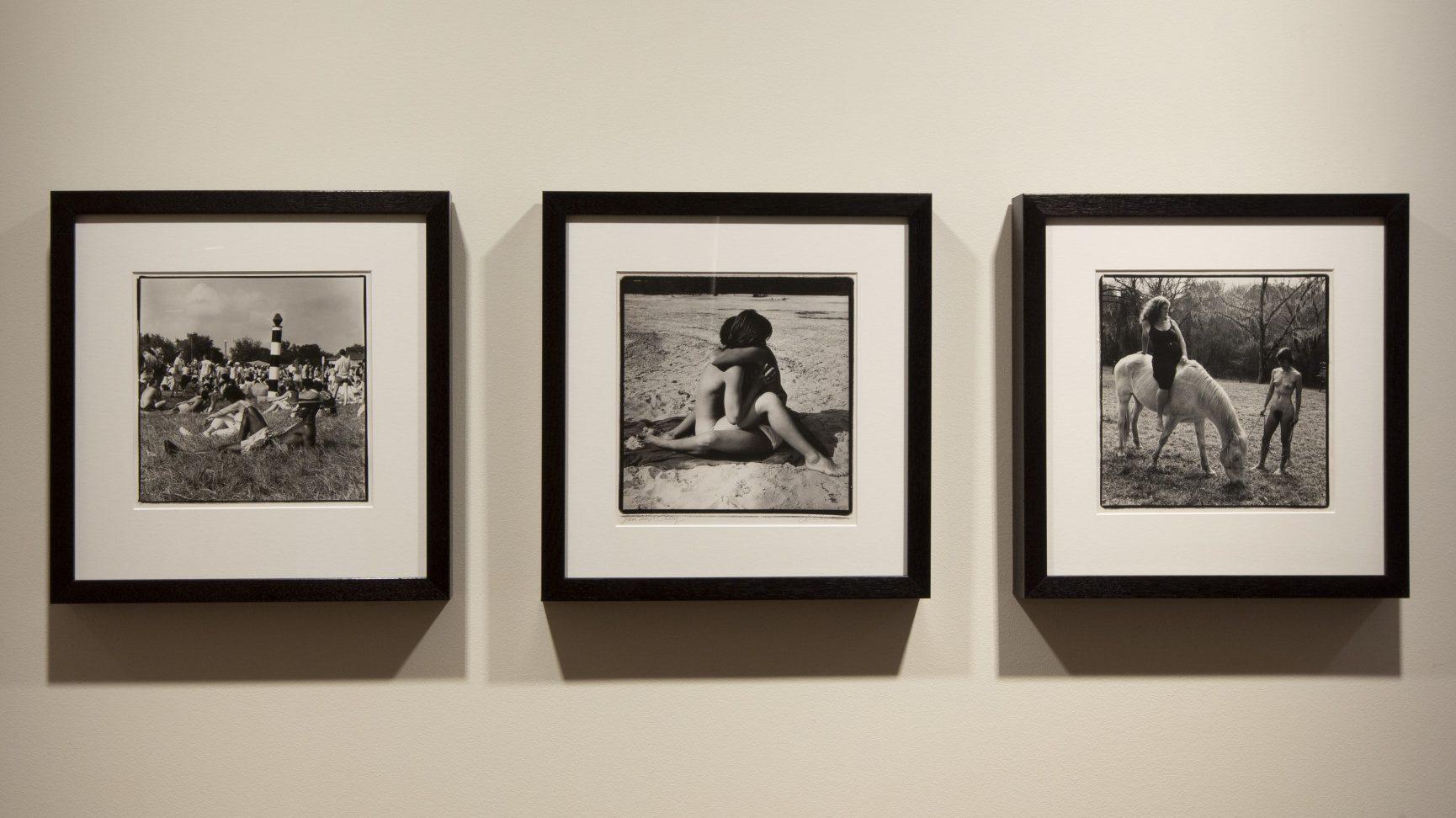 Three black-and-white framed photographs on a wall. The first shows a group of people sitting and standing in a grassy area. The second features an intimate beach scene. The third depicts a person with a horse in an outdoor setting.