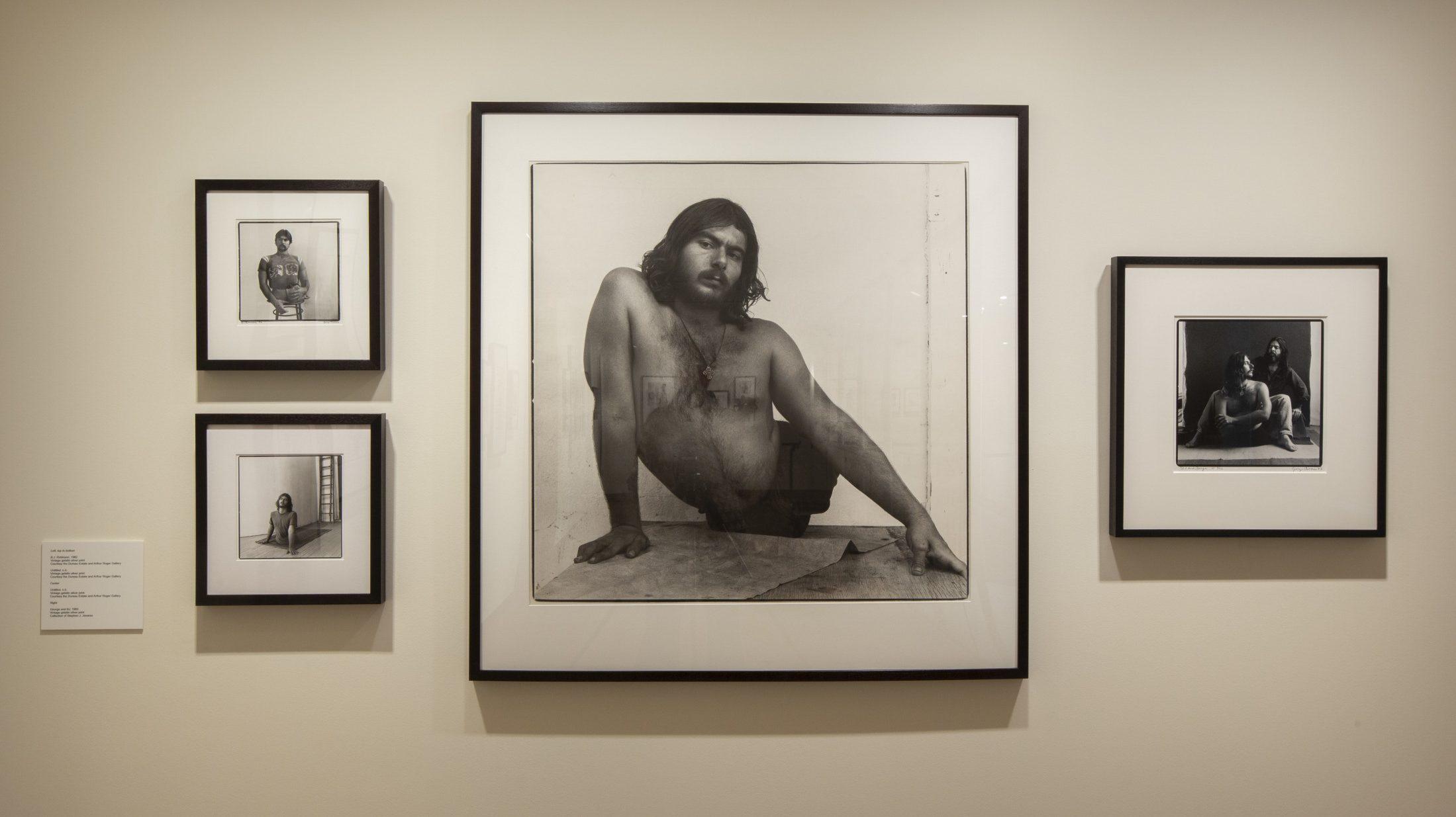 A gallery wall displaying four framed black and white photographs of a person posing in various seated positions. The largest photograph is centrally placed with three smaller ones surrounding it. The wall is off-white, creating a stark contrast with the frames.