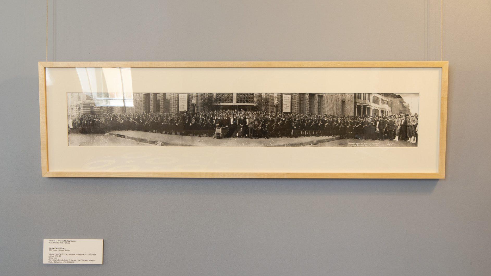 A framed panoramic black and white photo hangs on a gray wall. The photo depicts a large group of people gathered outside a building. A descriptive plaque is mounted below the frame on the wall.