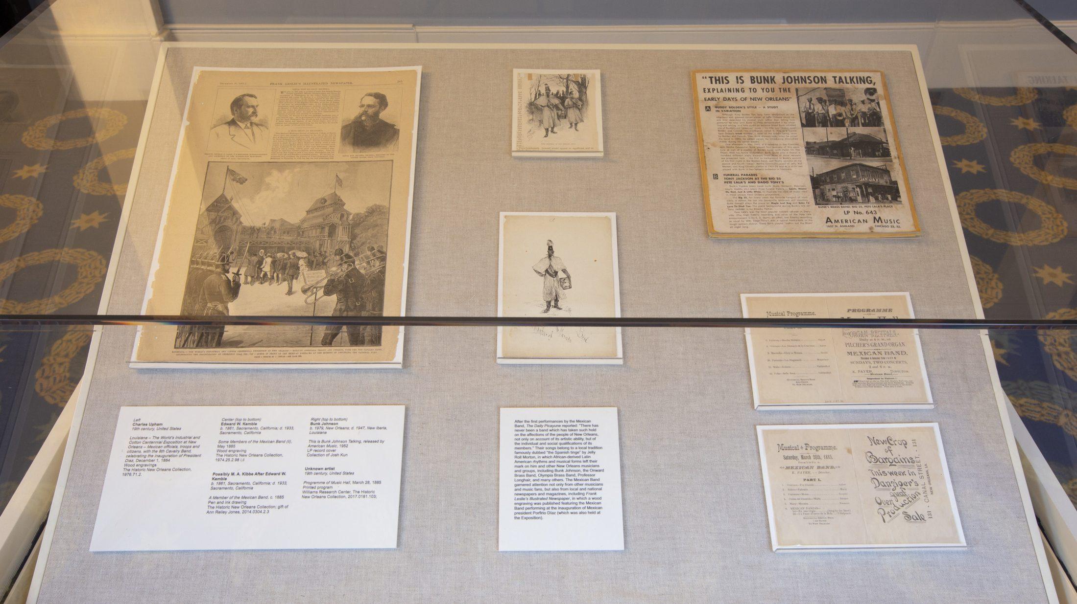 A display case containing historical documents, photographs, and newspaper clippings related to music history. Items are laid out on a cream-colored fabric background under glass.
