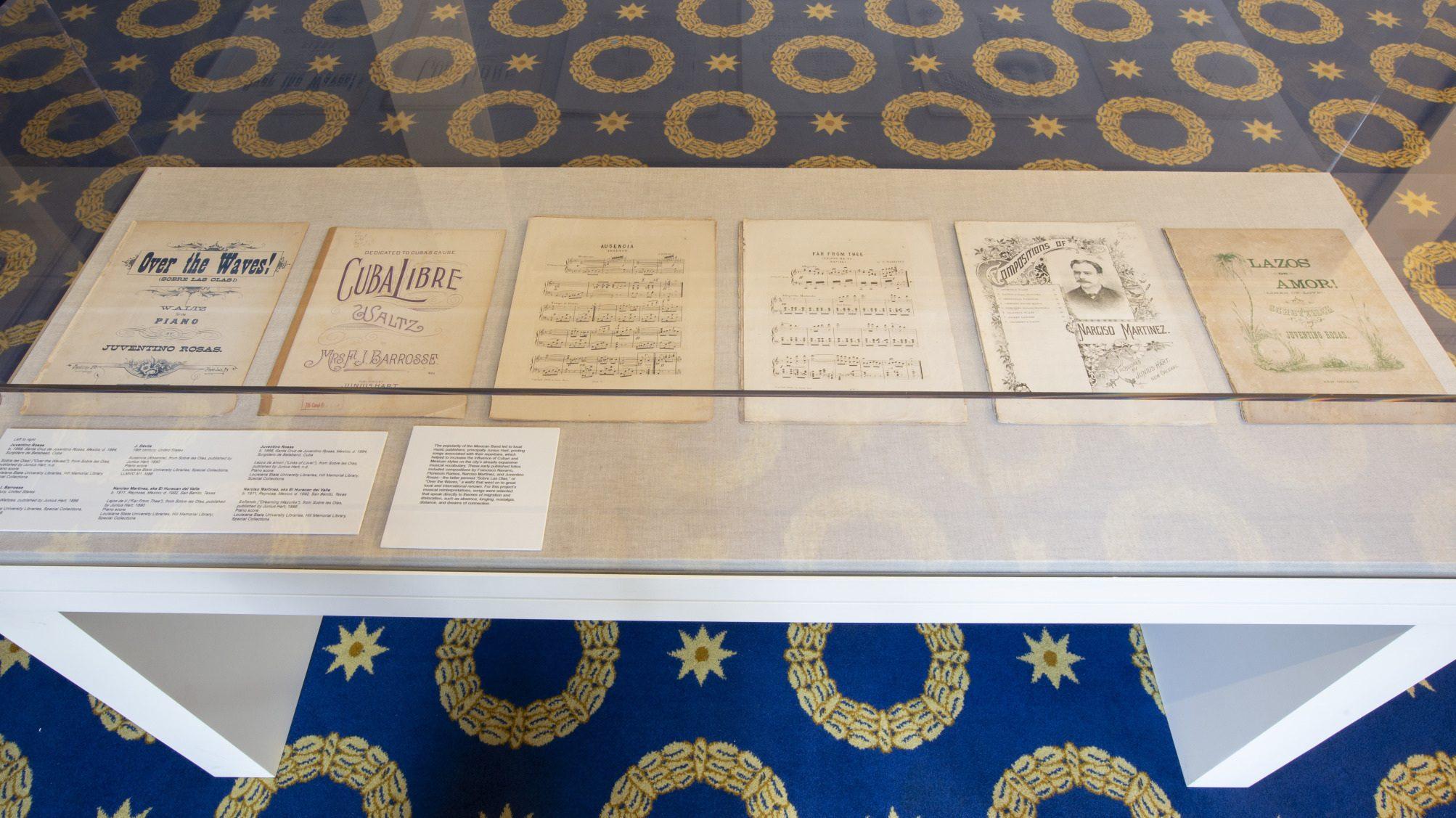 A display case showcases six music sheet covers laid out in a row. The sheets have various titles and designs. The case rests on a carpet with a blue background and gold circular patterns.