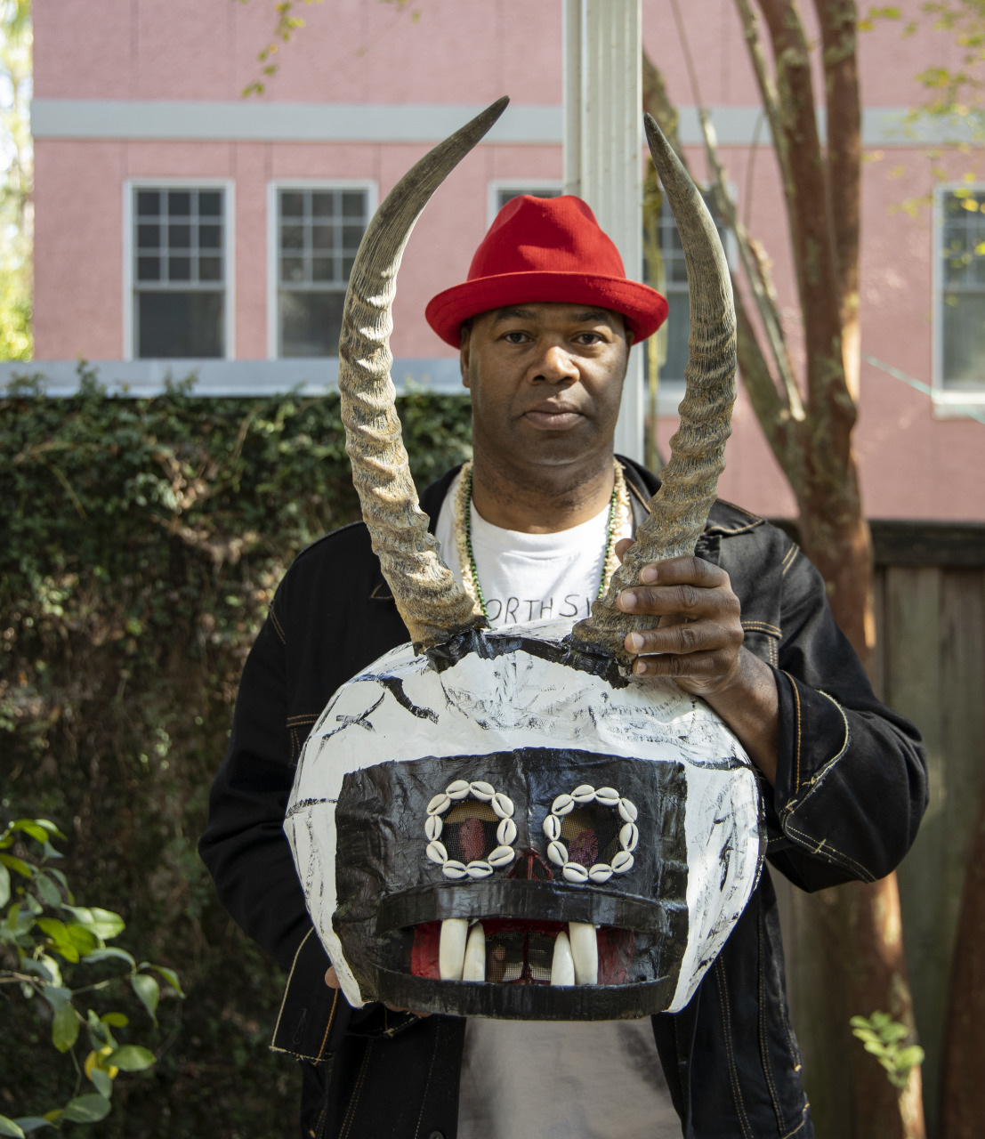A person stands outdoors holding a mask with large horns and tooth-like elements. The person is wearing a red hat, a black jacket, and a necklace. A pink building and trees are visible in the background.