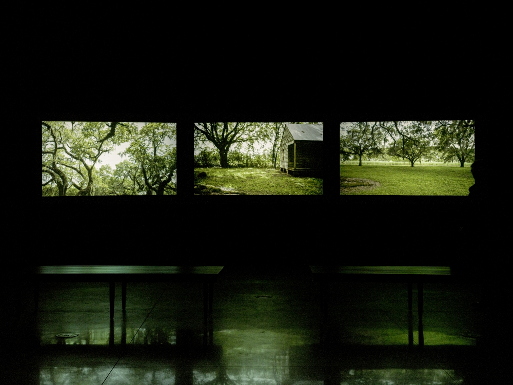 Three illuminated screens display images of a rural landscape featuring trees and a small cabin, set in a dark room with reflections on the shiny floor. Two benches are silhouetted in the foreground.