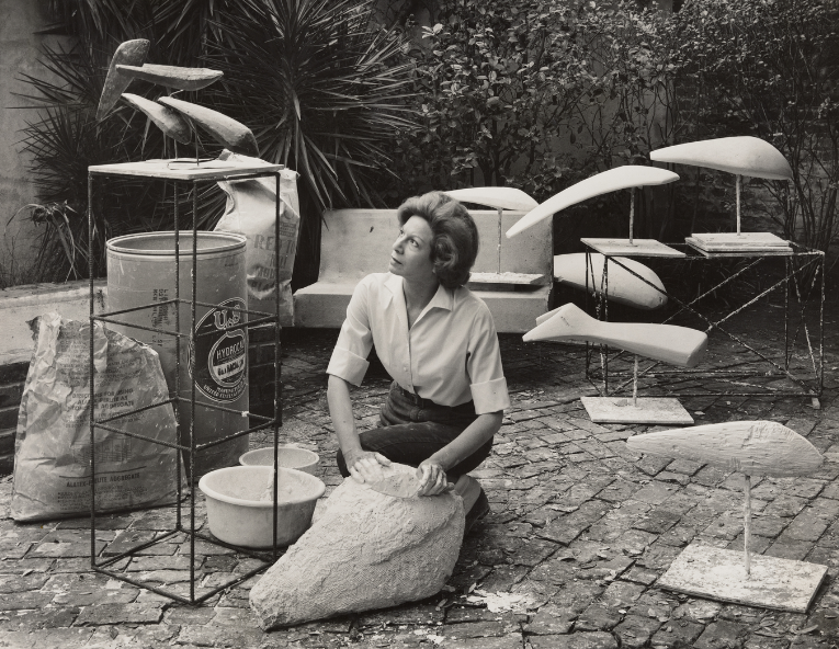 A woman kneels on a cobblestone surface, surrounded by abstract sculptures and art materials. She holds a large piece of plaster, with various sculpted forms and wire structures arranged around her in a garden setting.