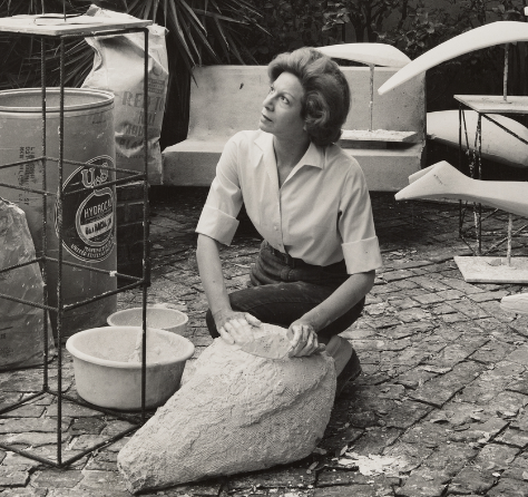A woman kneels on the ground, shaping a large piece of stone or clay. She is surrounded by art materials and sculptures in an outdoor setting with a brick floor and plants in the background.