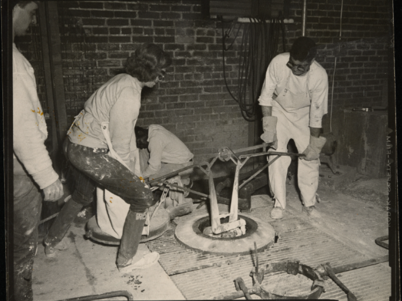 A group of people wearing protective clothing are operating metalwork equipment in an industrial setting with brick walls. Two are handling a large tool over a circular opening on the floor, possibly involved in a foundry process.