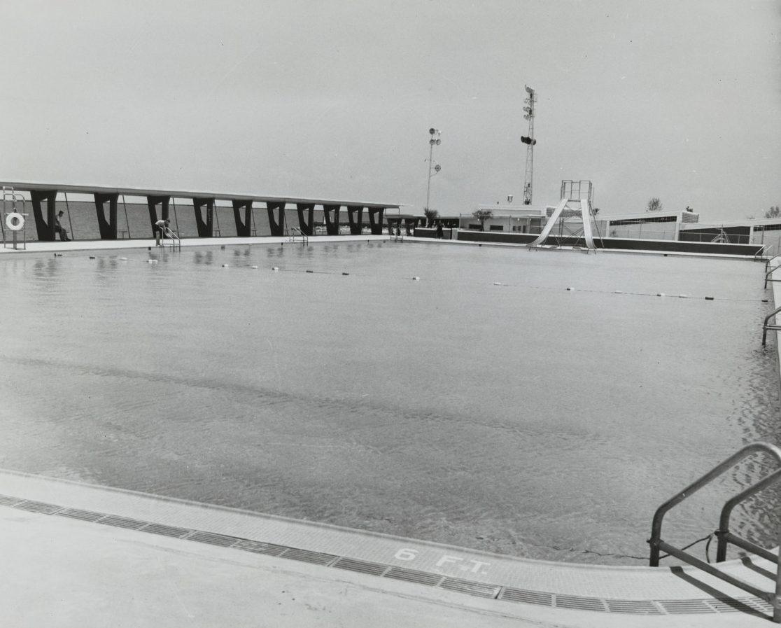 Black and white image of an outdoor swimming pool with a slide and marked lanes. The surrounding area includes a high fence and lifebuoys. The depth marker reads 6 FT near the pools edge. Overcast sky in the background.