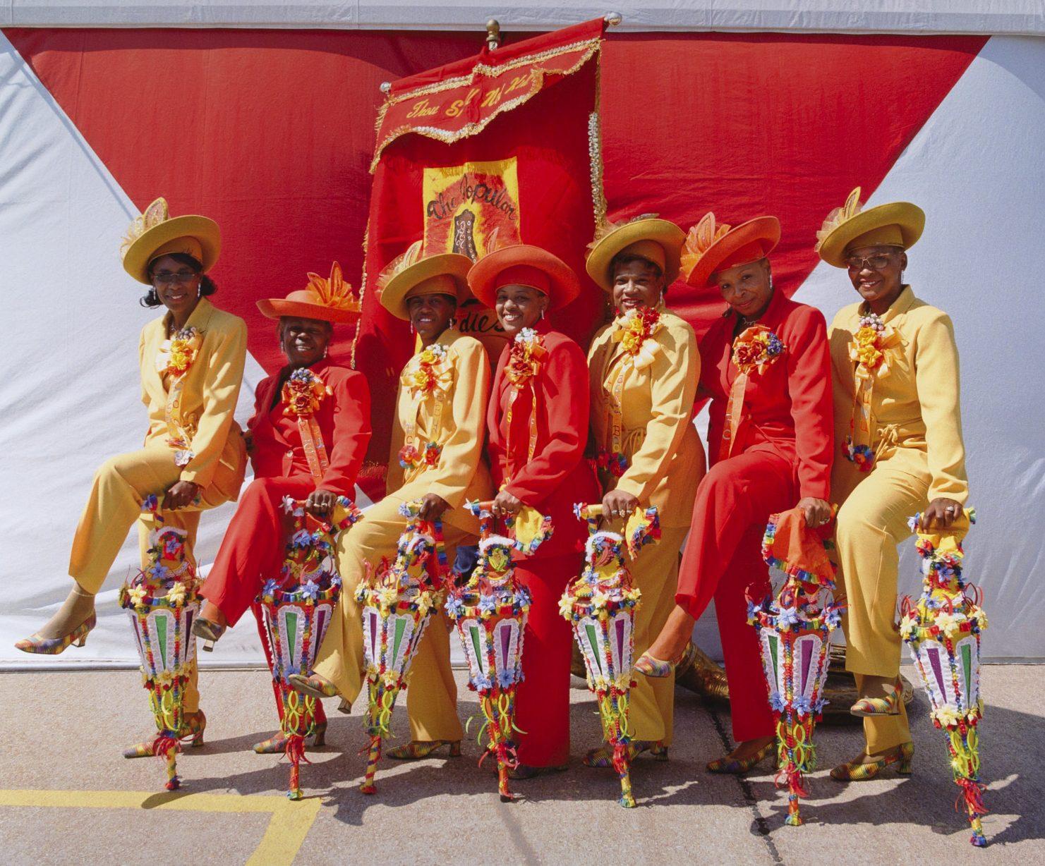 A group of seven people dressed in vibrant yellow and red outfits with matching hats pose with colorful, decorated umbrellas. They stand in front of a red and white banner, smiling for the photo.