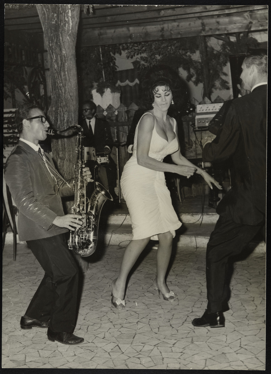 A lively black and white scene showing a woman in a white dress dancing energetically with a man in a suit. Another man plays the saxophone nearby. A band with a drummer is in the background on a stage. The setting appears to be an outdoor event.