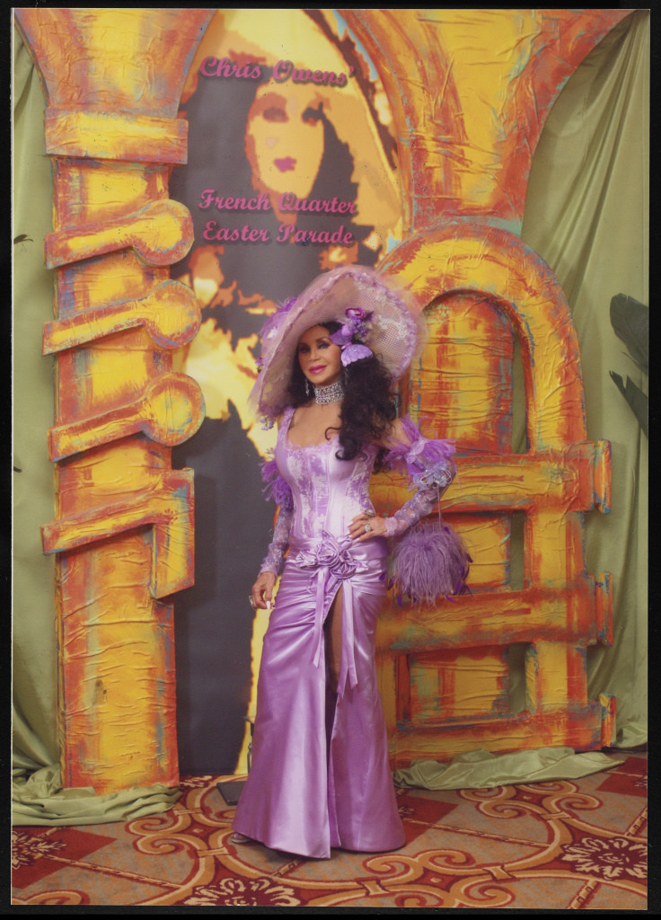 A person dressed in an elaborate purple outfit with a large hat, standing in front of a colorful backdrop featuring a vintage-style poster. The clothing includes ruffles and flowers, matching the vibrant setting.