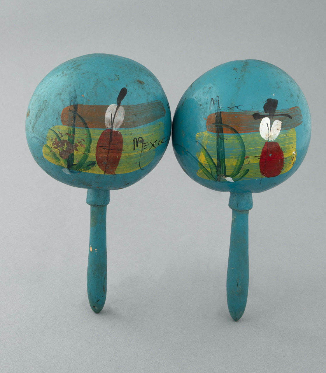 A pair of blue maracas with colorful painted designs, including abstract shapes and the word Mexico, set against a plain gray background.