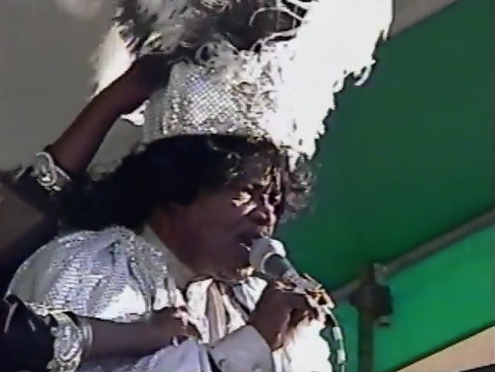 A person dressed in a sparkly silver outfit and headpiece sings into a microphone. They are standing in front of a green and white background, with a feathered detail on top of the headpiece.