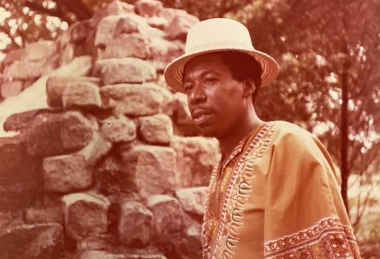 A person wearing a hat and an embroidered garment stands outdoors near a stone structure, with trees in the background. The image has a warm, sepia-like tone.