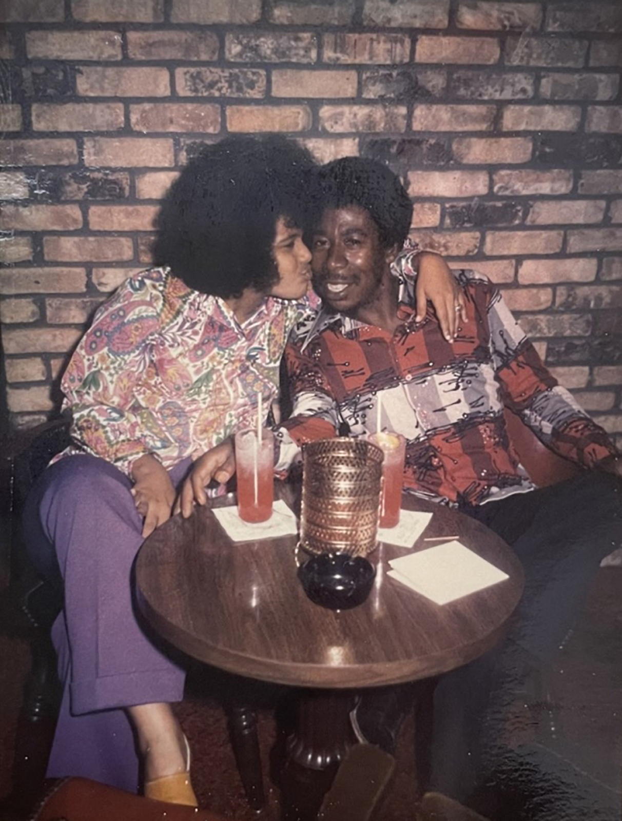 A couple sitting closely together in a cozy, dimly-lit setting. They are dressed in vibrant 1970s-style clothing, sharing smiles and drinks. The background features a brick wall, and a small round table holds two glasses and a decorative candle.
