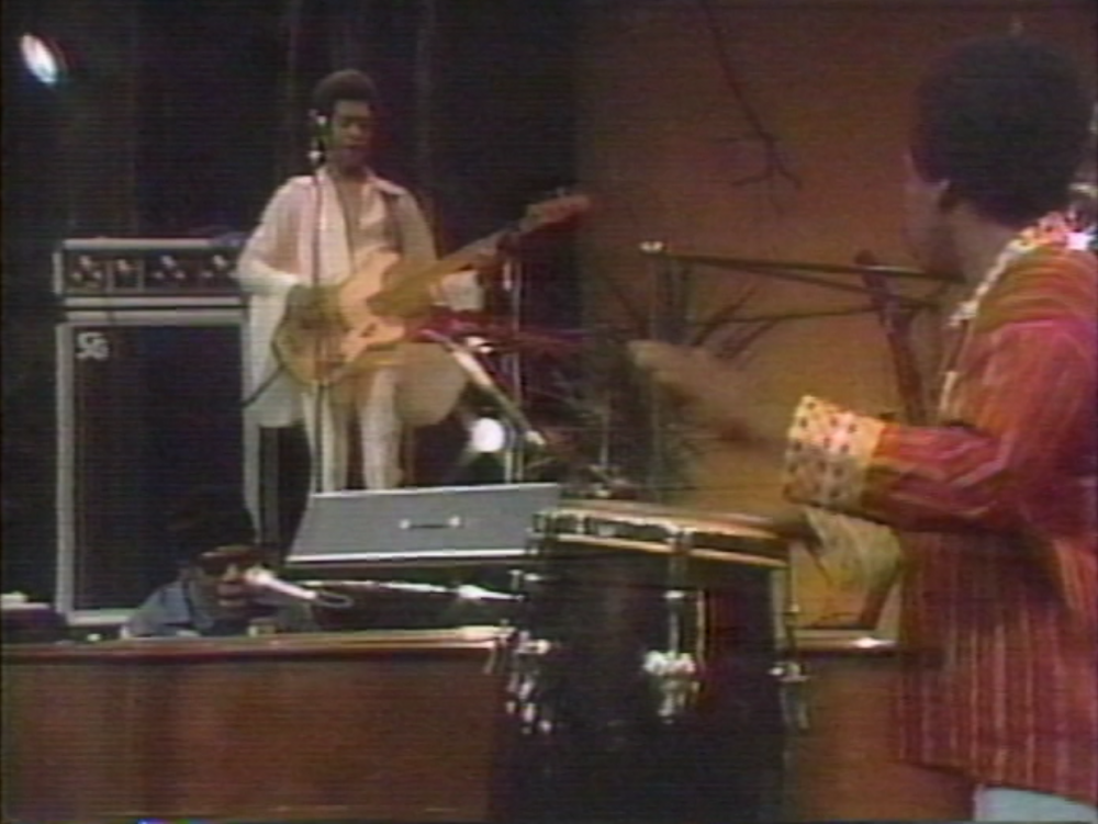 Musicians performing on stage with one playing a bass guitar and another on drums. A keyboardist is also visible, focusing on his instrument. The setting appears to be a live concert with musical equipment and instruments present.