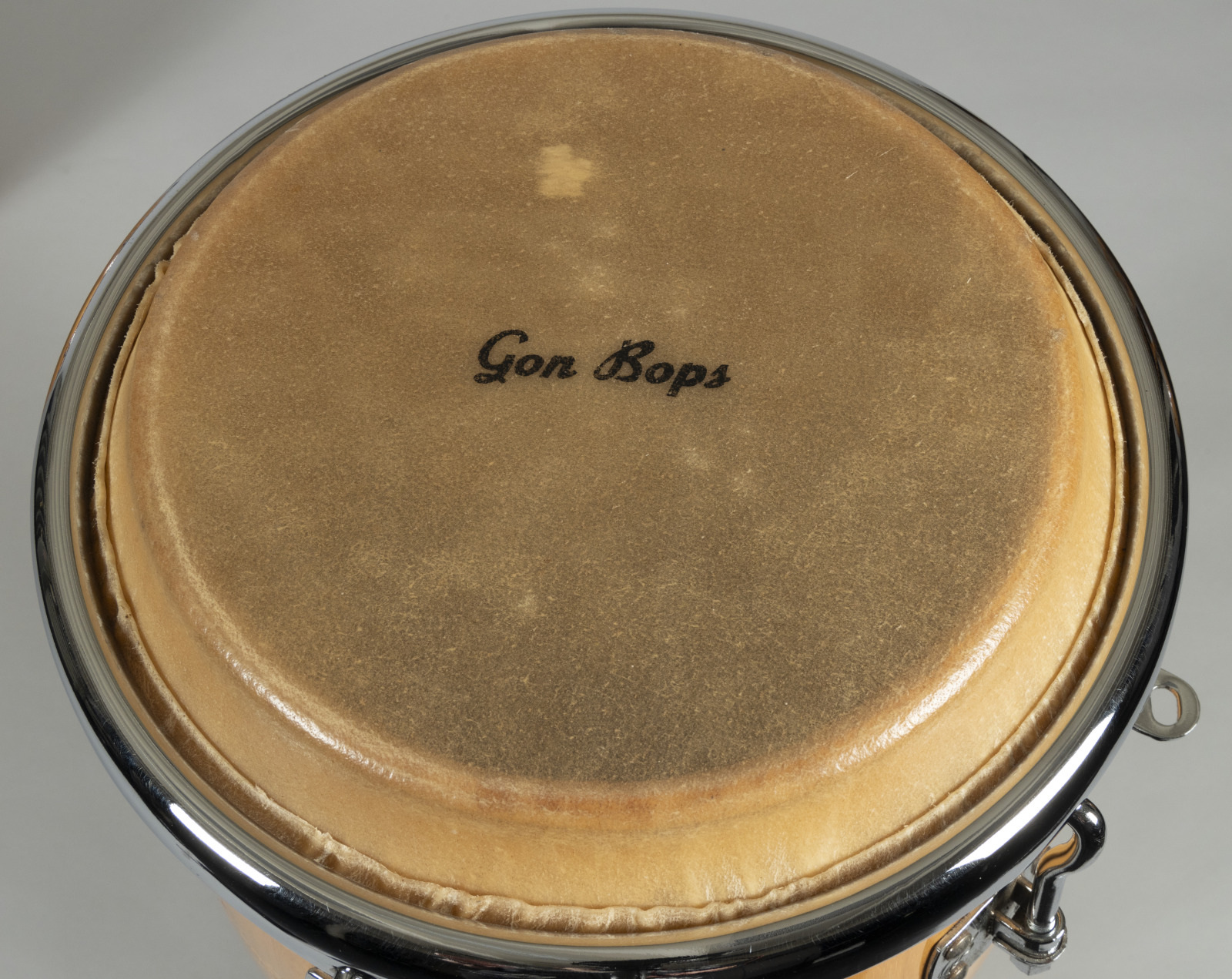 Close-up of a conga drum with Gon Bops written on the drumhead. The drum has a light brown wooden body and is surrounded by chrome hardware. The texture of the drumhead is visible, set against a neutral background.