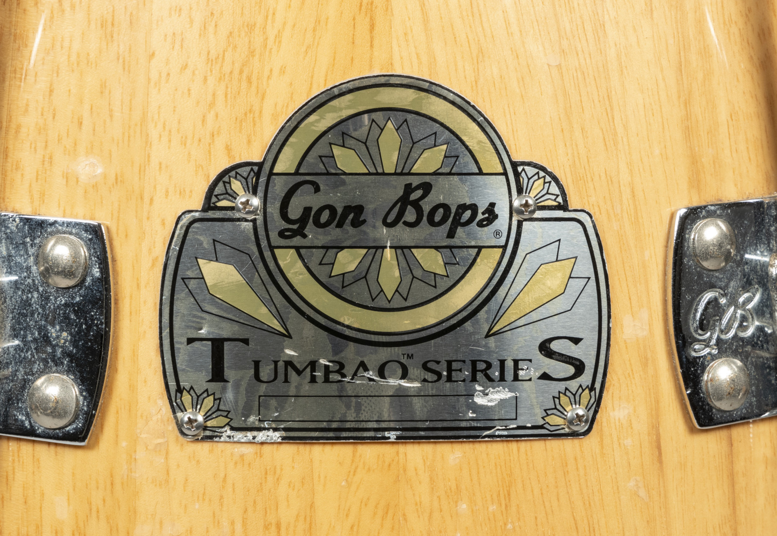 Close-up of a wooden drum with a metallic logo plate. The logo reads Gon Bops Tumbao Series with decorative floral motifs. Two shiny metal fixtures are visible on either side of the plate.