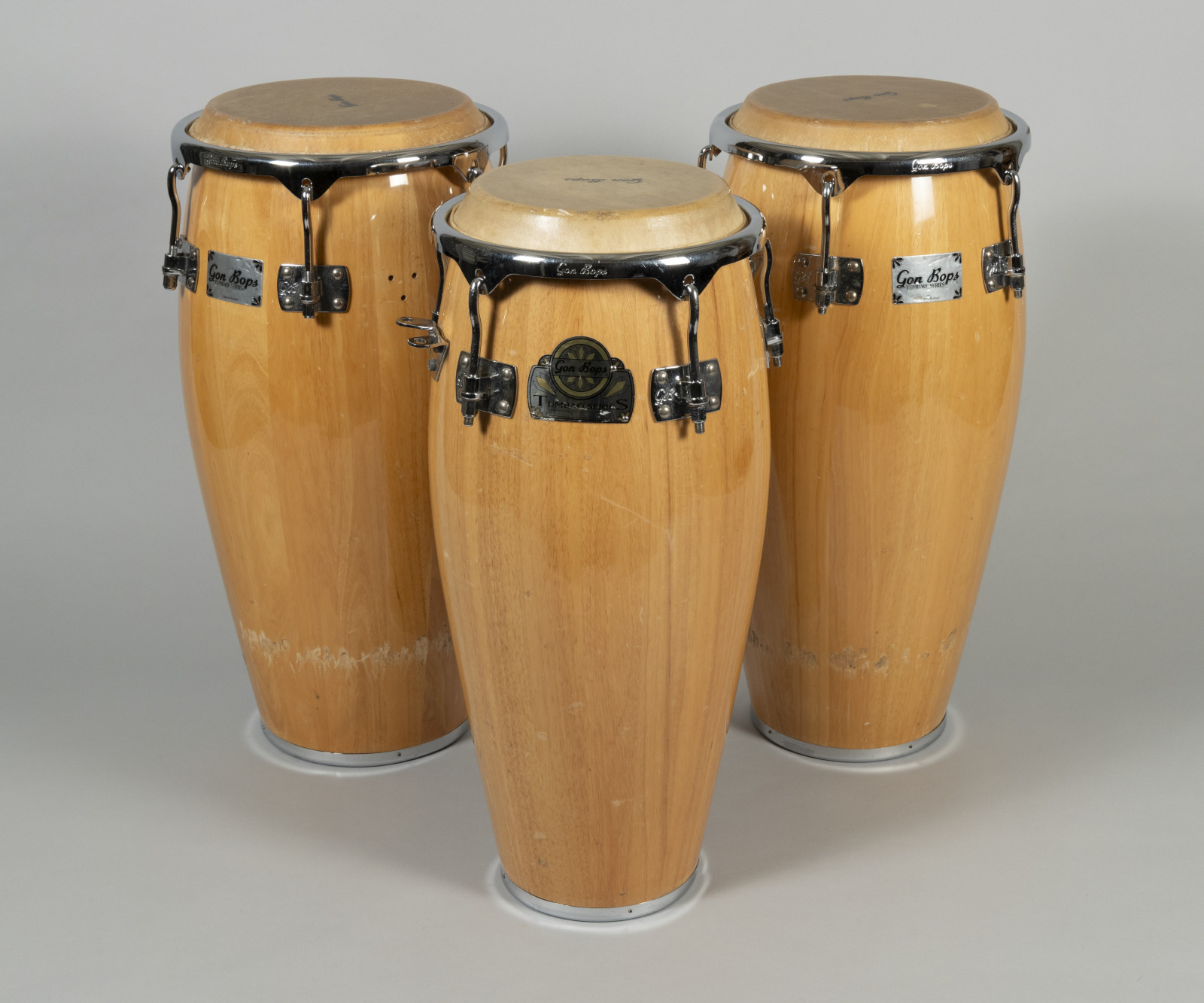 Three light brown conga drums are standing upright on a gray background. Each drum has a light wood finish with metal hardware at the top and bottom, and they are arranged in a triangular formation.