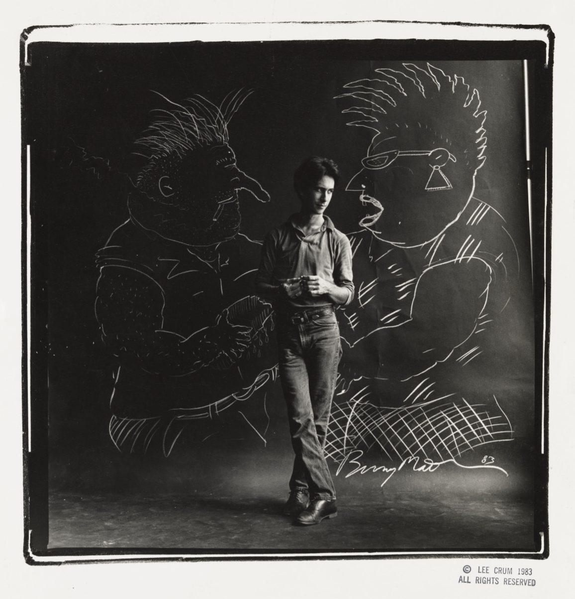 A man stands in front of a backdrop with two large chalk drawings of stylized figures wearing sunglasses. He is casually dressed, holding a cigarette, and the scene has a black and white, artistic tone.