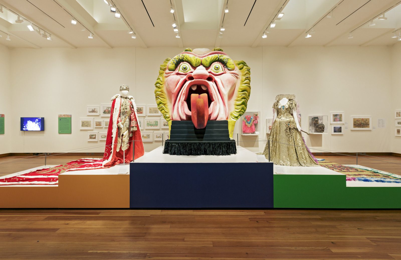 A museum exhibit featuring a large, colorful sculpture of a face with an open mouth and extended tongue. It is flanked by two elaborate costumes on mannequins. Artwork decorates the walls in the background.