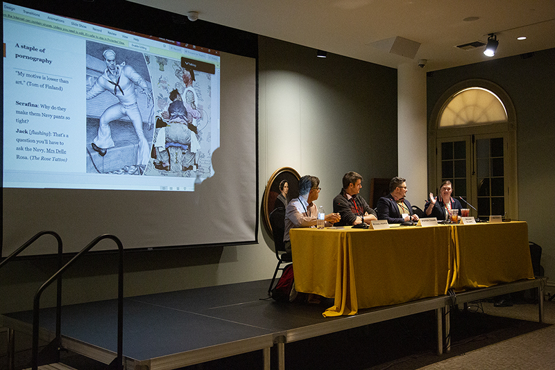 A panel of five people seated at a table on a stage. A projection screen displays an artwork and text. The room is dimly lit, and the audience is not visible.