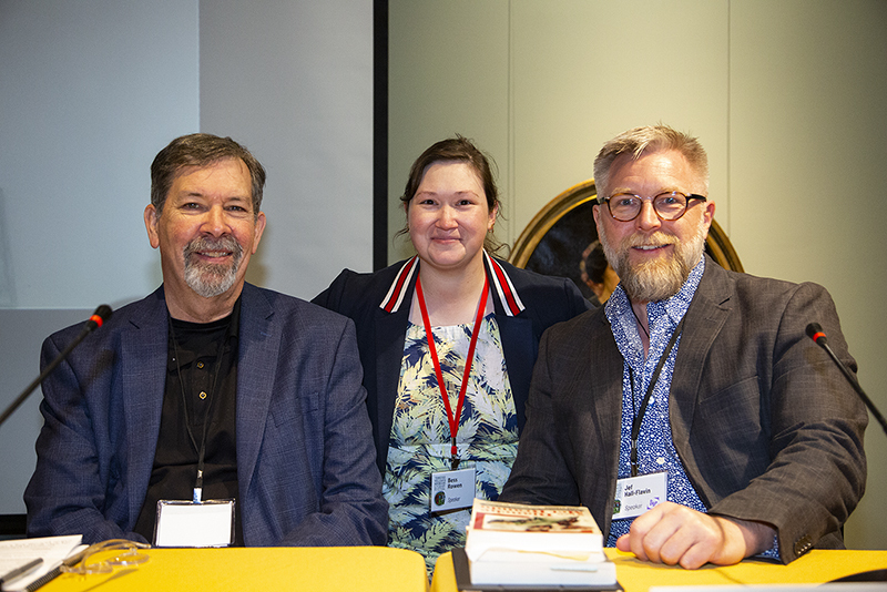 Three people pose together, smiling, at a conference. They wear name tags and casual business attire. One person in the middle stands behind a yellow table, while the others sit on either side. Books and microphones are in front of them.