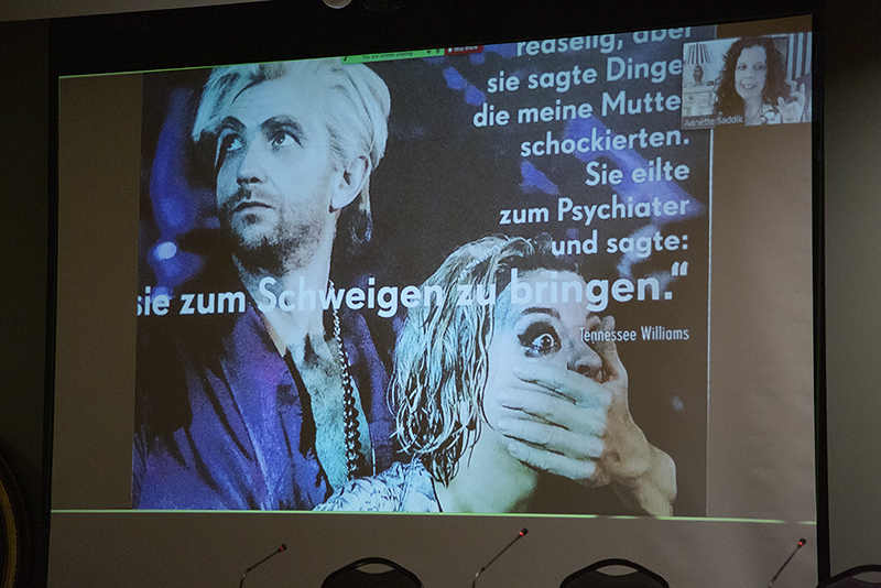 A large screen displays an image of a man with his hand over a womans mouth, accompanied by German text and the name Tennessee Williams. A small video call window shows a smiling woman in the top right corner.