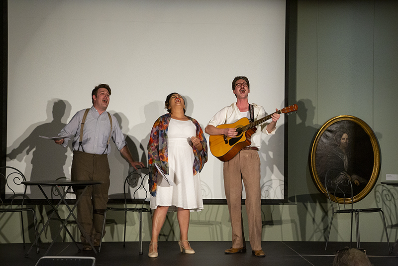 Three performers on stage sing energetically. One plays an acoustic guitar, while two hold papers. A portrait of a woman in an oval frame is visible on the wall. They are in front of a blank screen, standing by metal chairs.