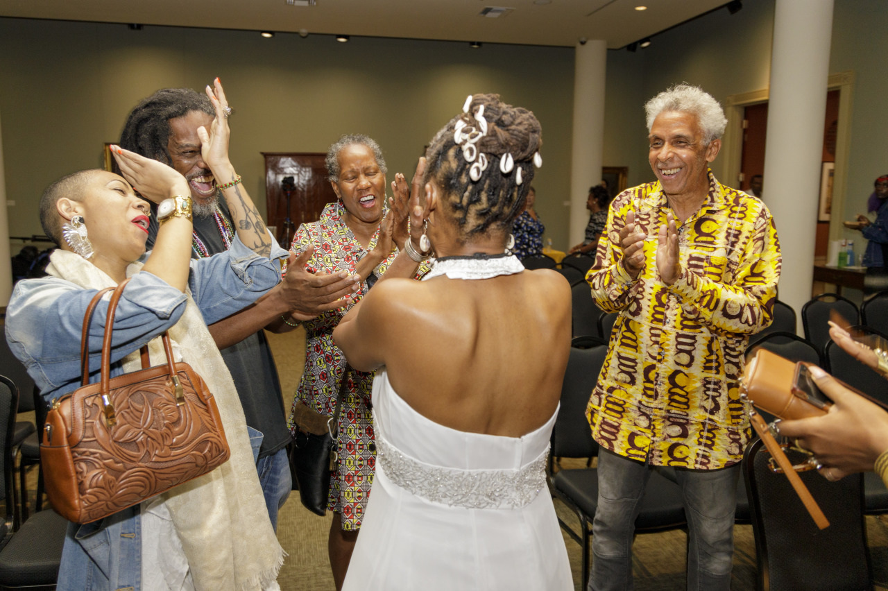 A group of people joyfully celebrating with a woman in a white dress at the center. They are smiling, clapping, and appear to be in a festive mood, set in a room with chairs and columns.