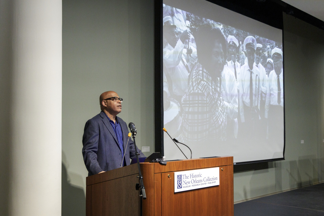 A man stands at a podium speaking, with a black and white historical photo projected in the background. The venue is labelled The Historic New Orleans Collection.