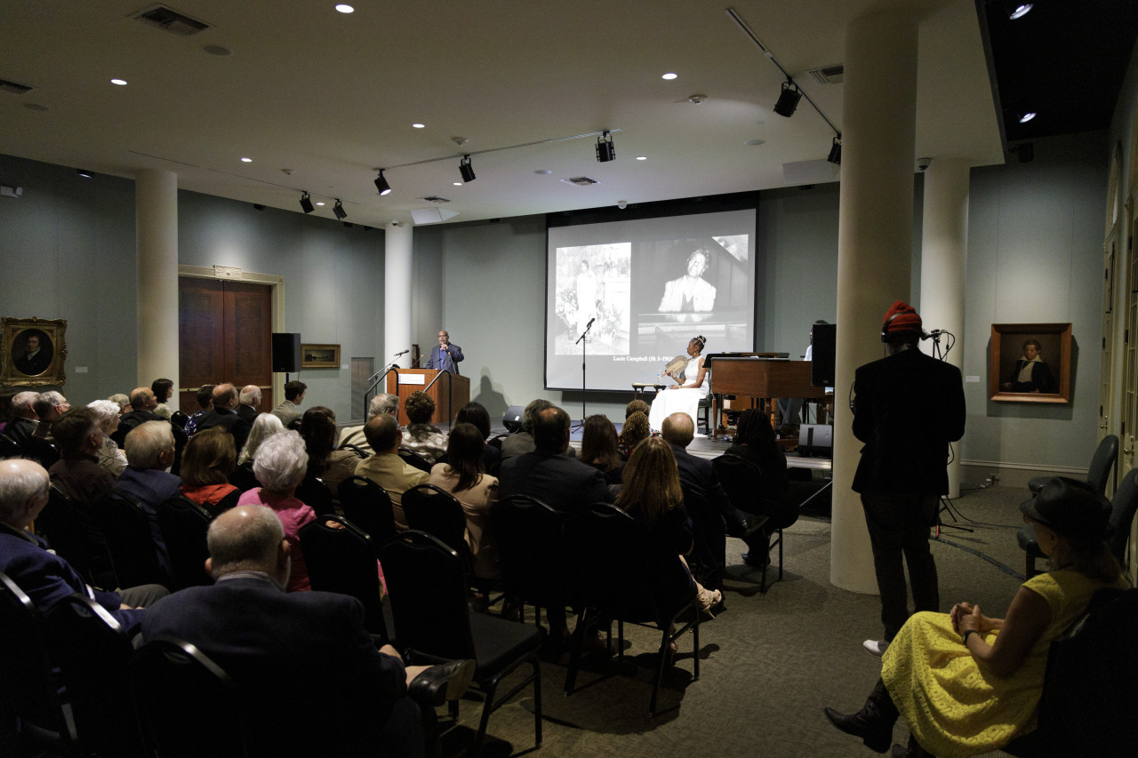 A speaker presents a slideshow in a dimly lit auditorium filled with seated attendees. The audience watches intently, and portraits hang on the walls. A person stands at the podium, while another sits on stage next to a piano.